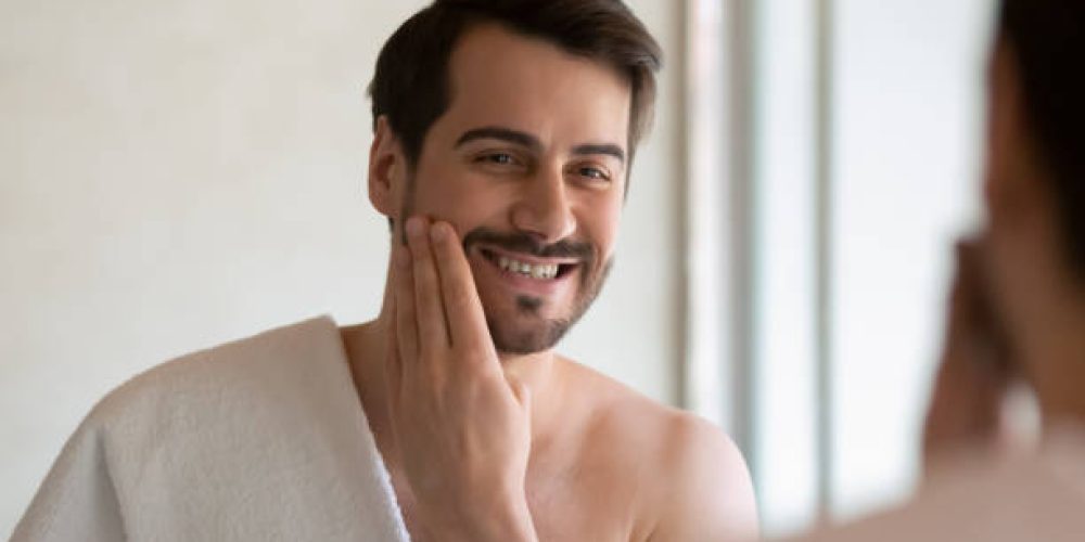 Smiling Caucasian millennial man in towel look in mirror apply face lotion after shaving in home bathroom, happy young male do daily skincare facial routine treatment in bath, hygiene concept